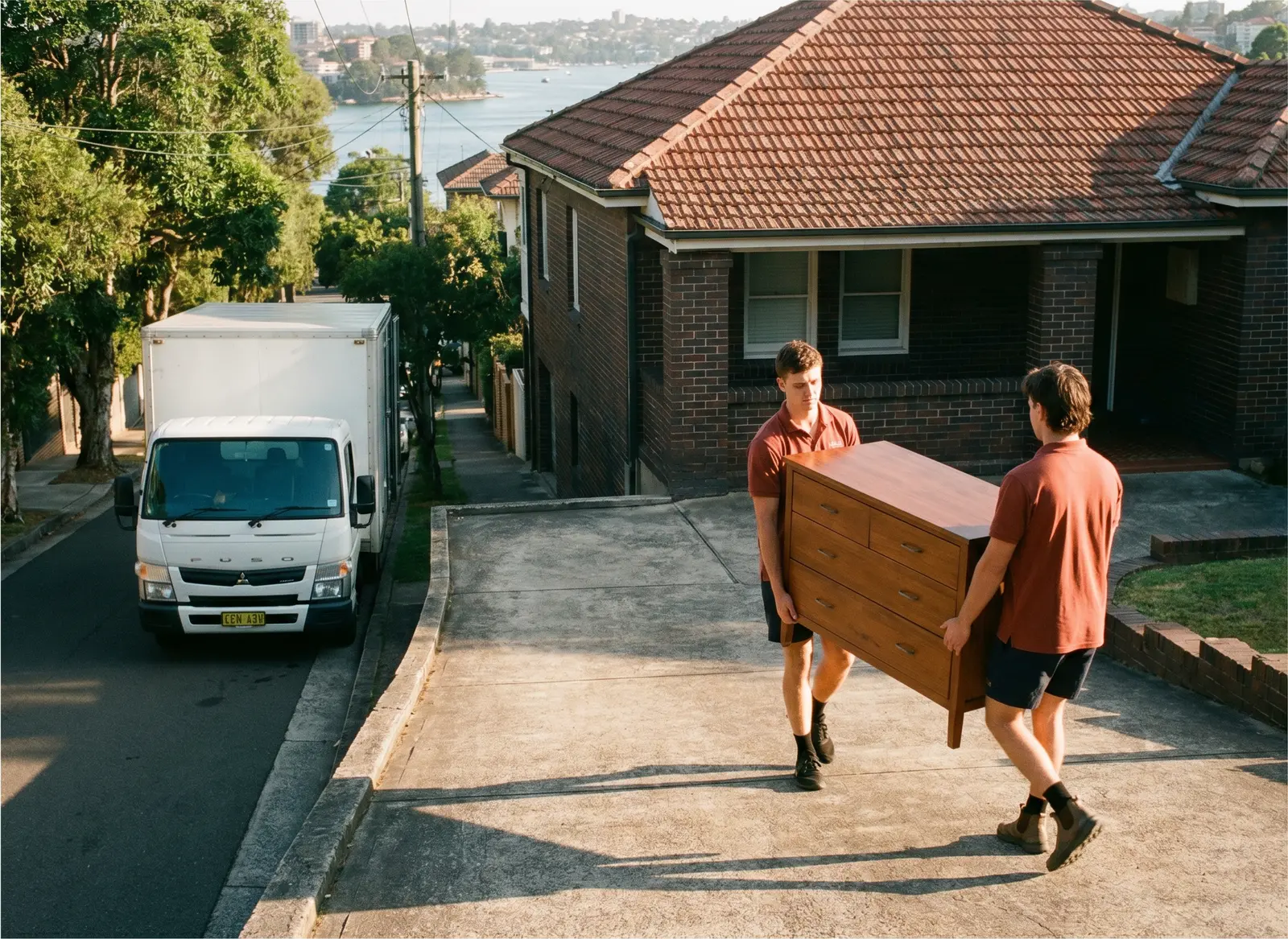 Two removalists carrying a timber chest of drawers up a steep concrete driveway, truck at the kerb below